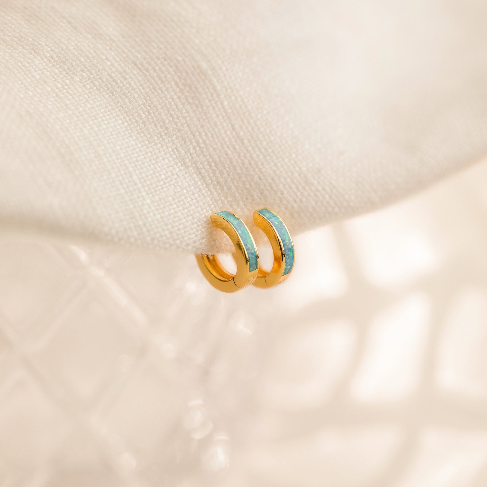 Two Ocean Opal Inlay Huggies rest on soft fabric—perfect as delicate wedding jewelry or a subtle accent alongside favorites like the Light Blue Opal Huggie Hoops.