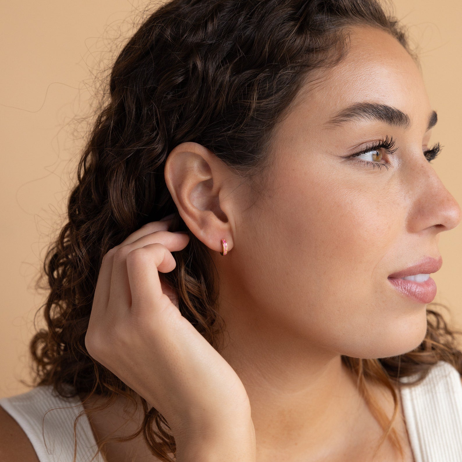 A woman with curly hair touches her ear, highlighting the elegant Magenta Opal Inlay Huggies.
