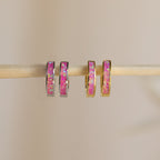 Two pairs of Magenta Opal Inlay Huggies, featuring pink and iridescent inlays, hang on a wooden rod. These earrings add a subtle pop of color to any look.