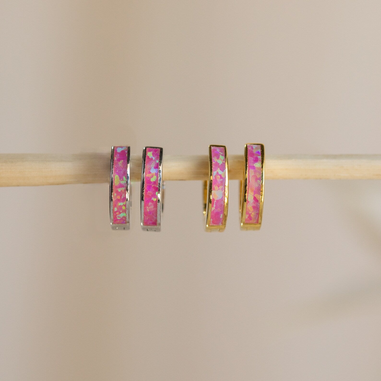 Two pairs of Magenta Opal Inlay Huggies, featuring pink and iridescent inlays, hang on a wooden rod. These earrings add a subtle pop of color to any look.
