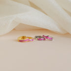 Two pairs of Magenta Opal Inlay Huggies, in gold and silver finishes, are displayed on a soft cream fabric background.