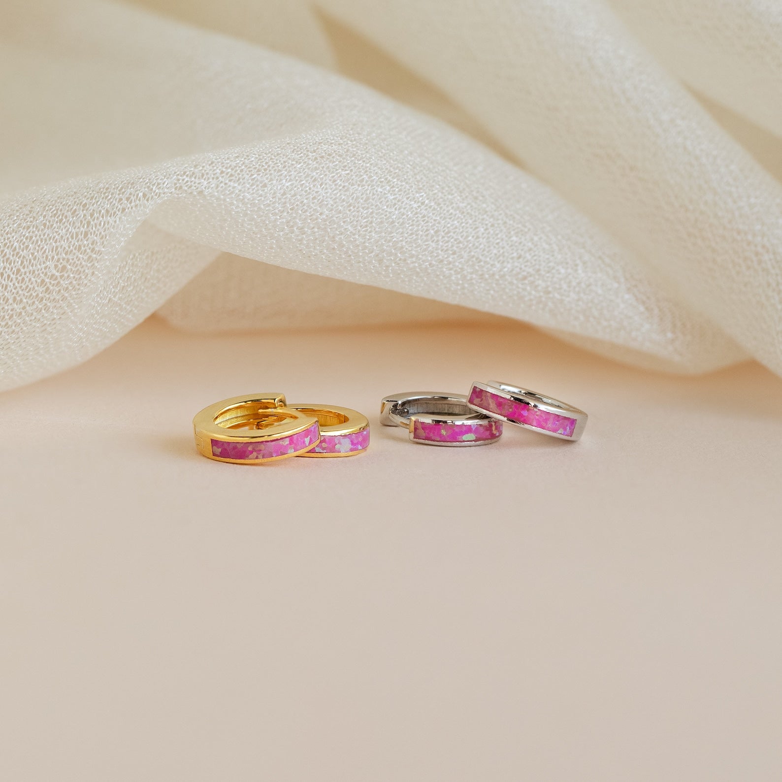 Two pairs of Magenta Opal Inlay Huggies, in gold and silver finishes, are displayed on a soft cream fabric background.