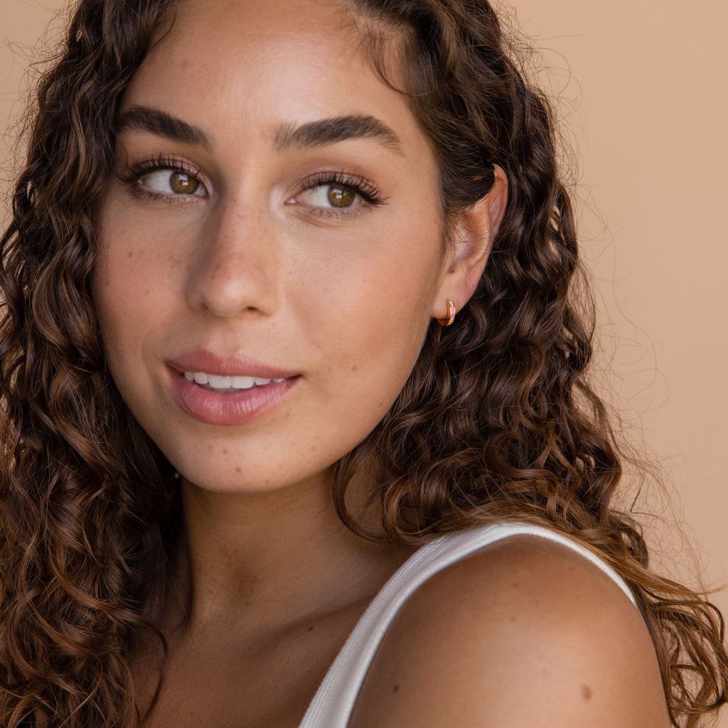 Woman with long curly hair wearing a white top and the Pink Opal Inlay Huggies, looking to the side against a beige background.
