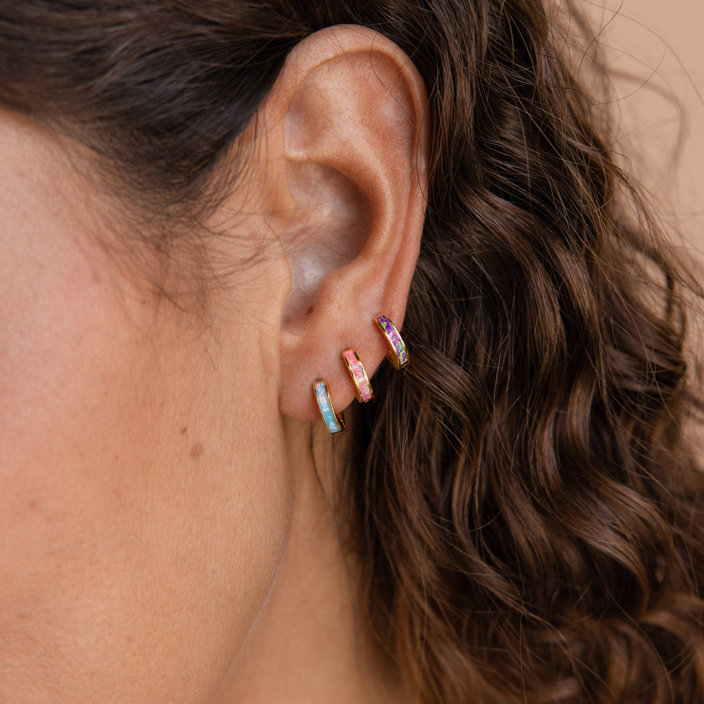 A close-up of a person's ear with three small, colorful hoop earrings—including the Pink Opal Inlay Huggies—adorns their curly brown hair.