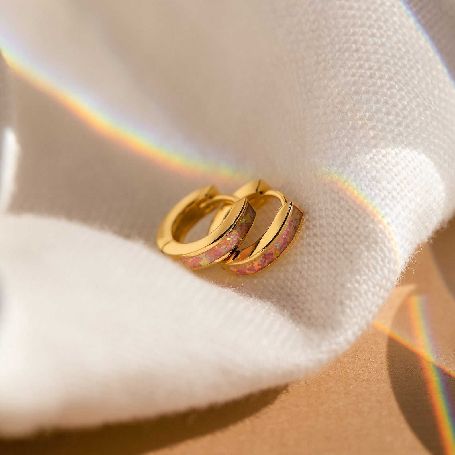 Pink Opal Inlay Huggies rest on soft white fabric, illuminated by rainbow light.