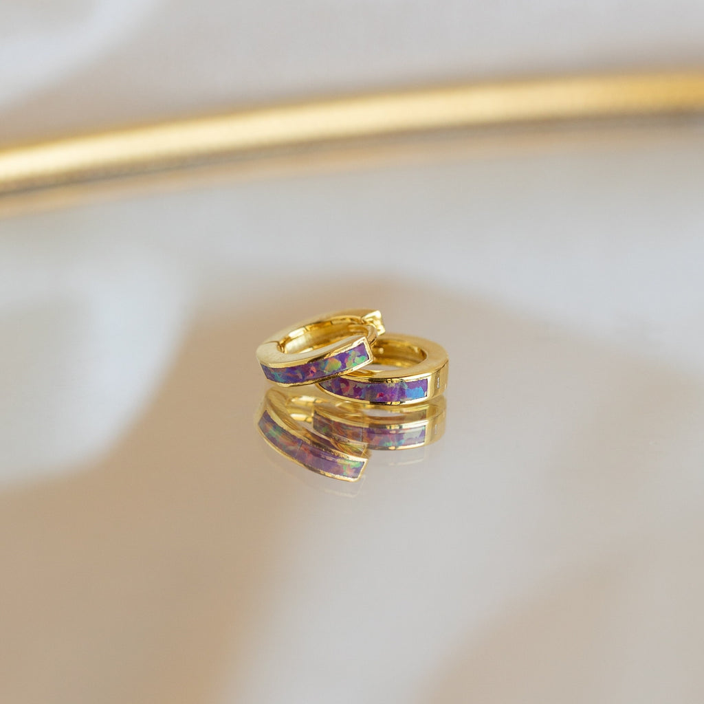 Lavender Opal Inlay Huggies with purple and red accents, featuring a subtle opal shimmer, rest elegantly on a reflective surface.