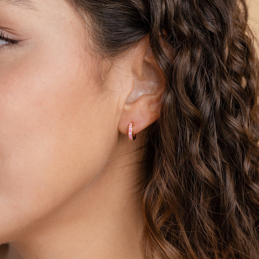 Close-up of a woman's ear with curly brown hair, wearing Magenta Opal Inlay Huggies—small pink hoop earrings that add a vibrant touch.