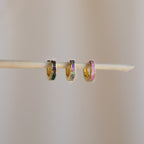 Three Magenta Opal Inlay Huggies, small gold hoops with colorful inlays, hang delicately on a thin wooden stick against a plain background.