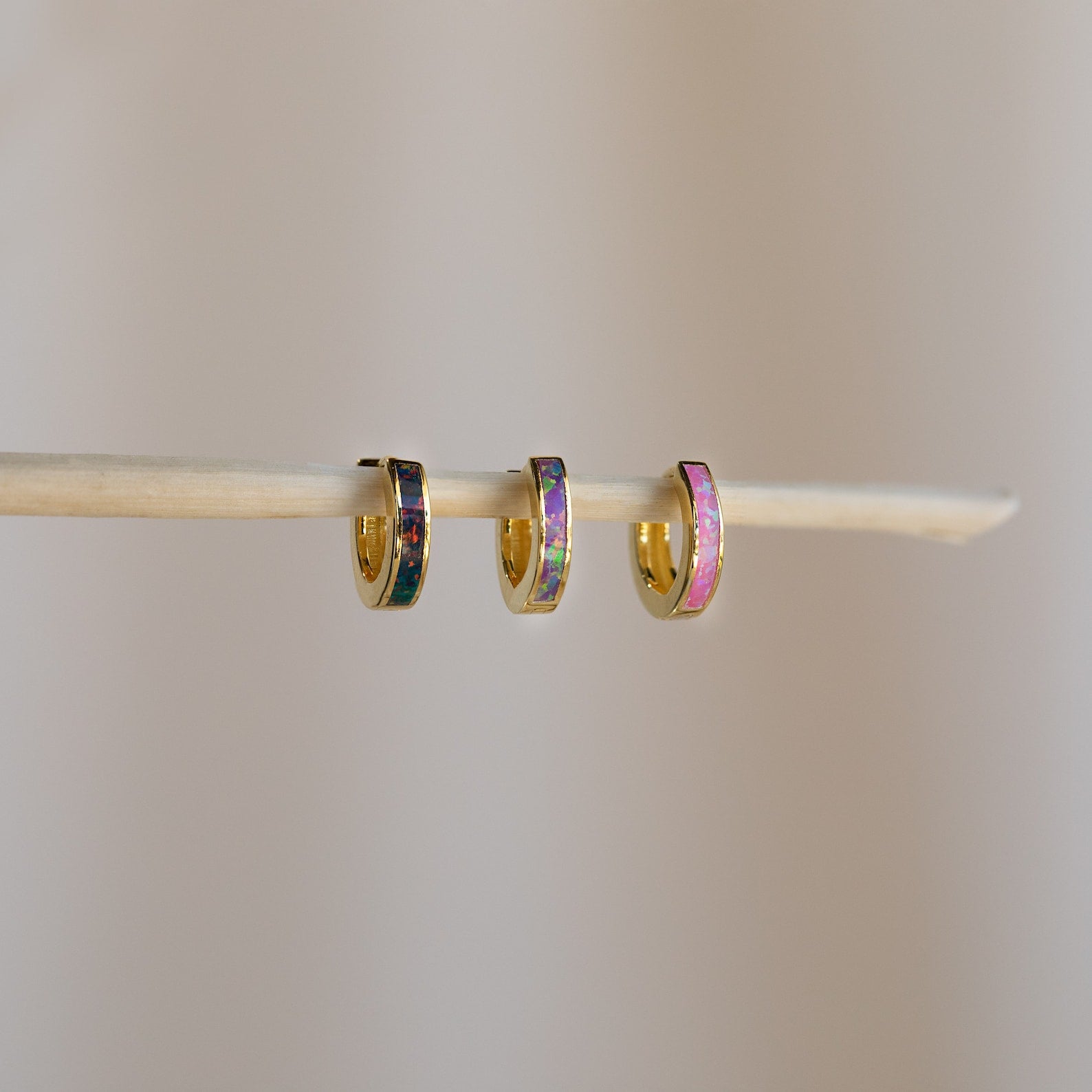 Three Magenta Opal Inlay Huggies, small gold hoops with colorful inlays, hang delicately on a thin wooden stick against a plain background.