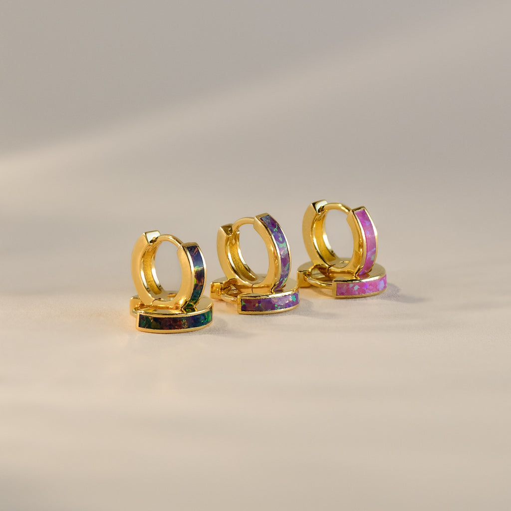 The Lavender Opal Inlay Huggies, a set of three minimalist hoop earrings with colorful iridescent accents, including a purple opal shimmer, are displayed on a soft beige background.