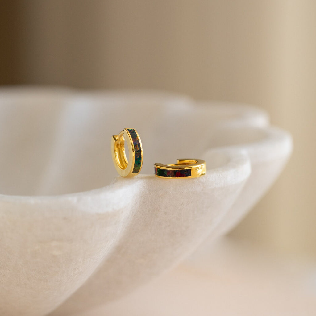 The Black Opal Inlay Huggies, two small gold hoop earrings with dark opal inlay, rest on the edge of a white, scalloped dish.