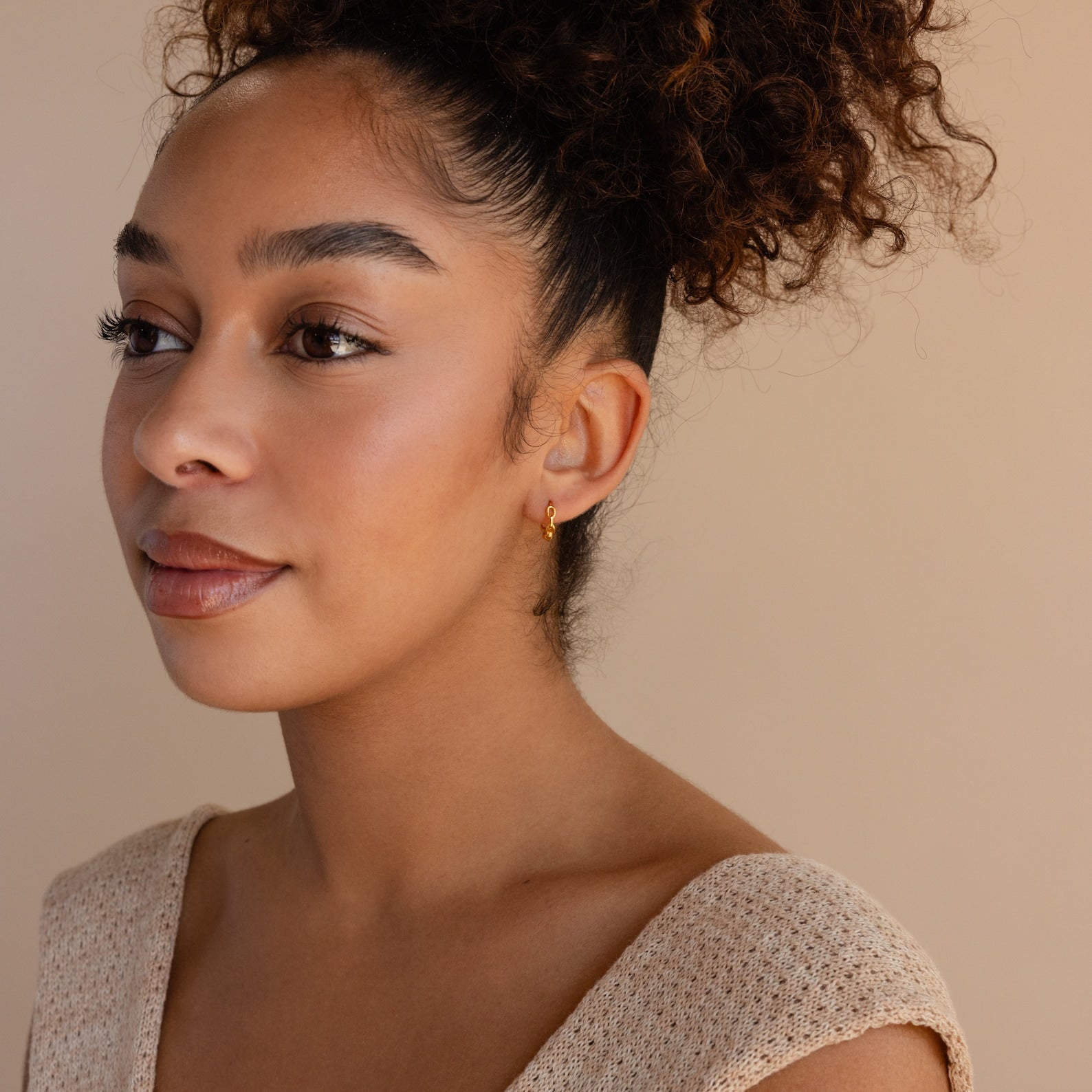 Woman with curly hair in a bun, wearing a beige top and an earring stack featuring Link Huggies and dainty chain earrings, looking to the left.