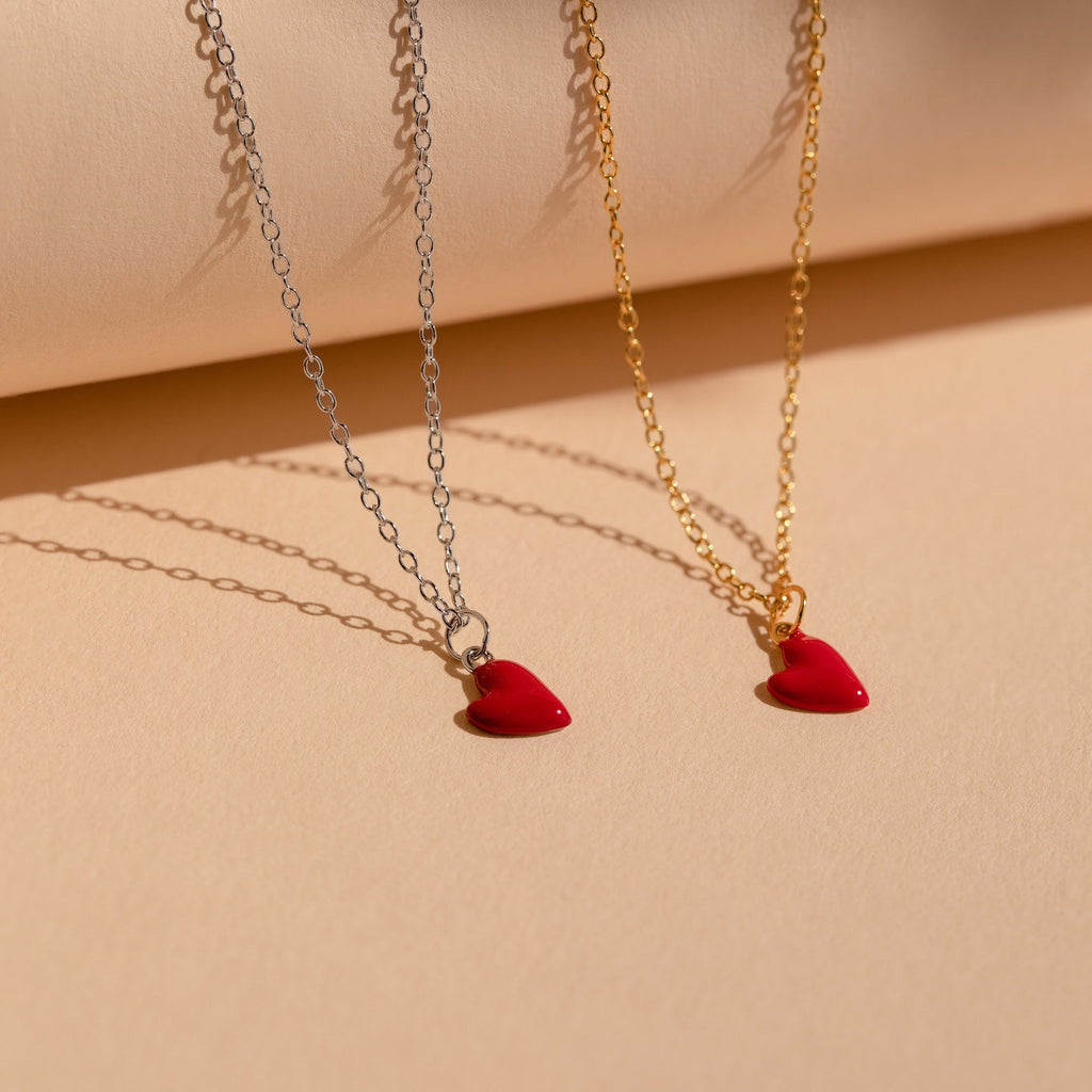 Two Little Red Heart Necklaces, one with a silver chain and one with a gold chain, are displayed on a beige background—an elegant choice for anyone loving the Little Red Heart Necklace trend.