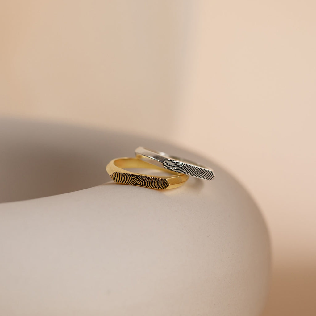The Thin Fingerprint Signet Ring, available in gold and silver with unique fingerprint designs, is displayed on a smooth beige surface—perfect as a promise ring or personalized gift for someone special.