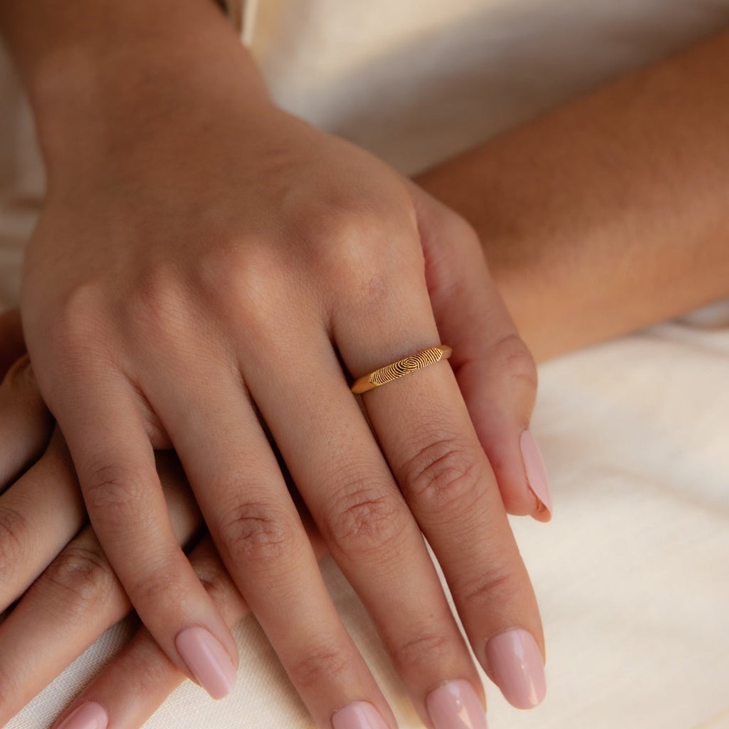 A hand with pale pink nails wears the Thin Fingerprint Signet Ring on the ring finger against a neutral fabric background.