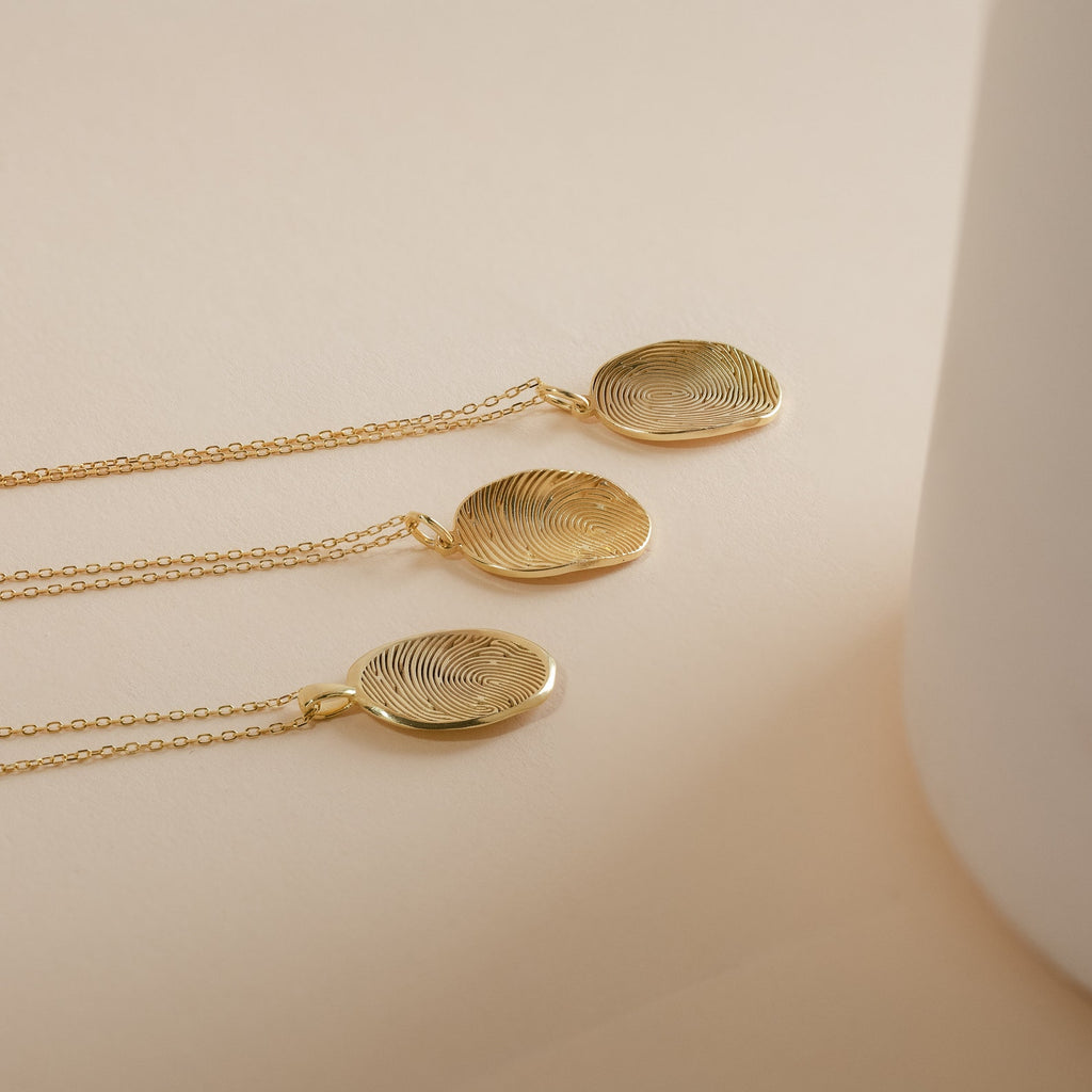 Three gold Custom Fingerprint Necklaces with round pendants rest on a beige surface beside a white object. Each personalized necklace captures your unique touch, making it a meaningful keepsake.