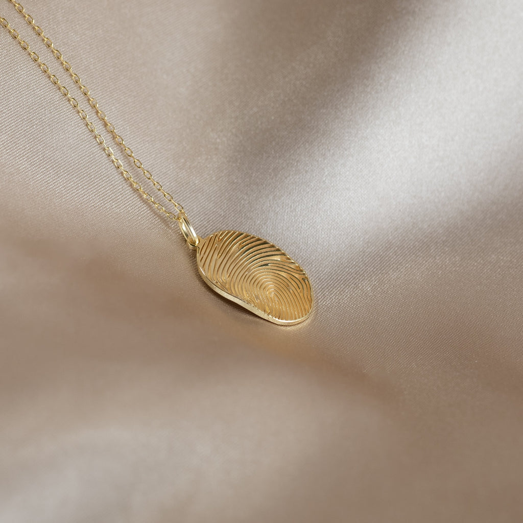 The Custom Fingerprint Necklace features an oval pendant with your unique fingerprint, displayed on smooth, light beige satin. A timeless personalized jewelry piece, ideal for cherishing treasured memories.