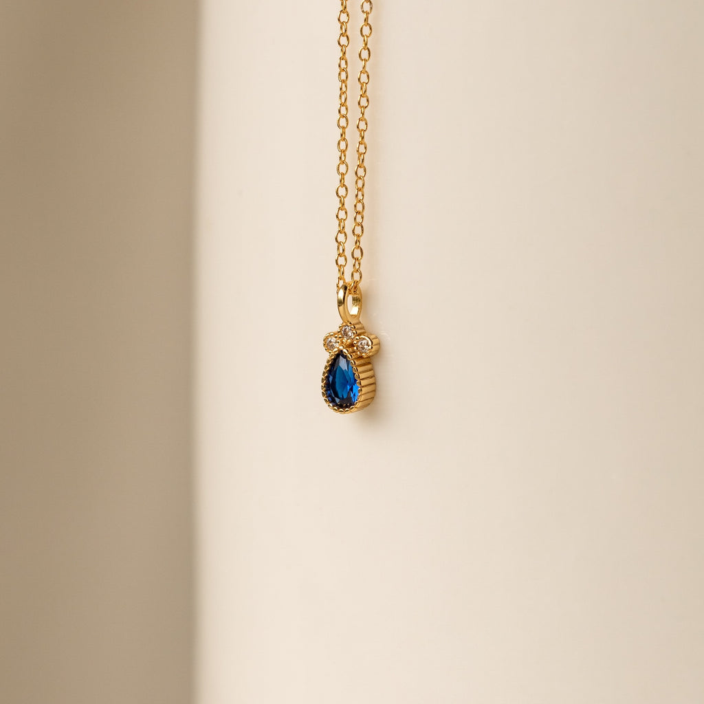 The Lorelei Sapphire Teardrop Necklace features a gold chain with a sapphire teardrop pendant and clear stones, creating an elegant, timeless accessory perfect for any outfit.