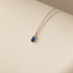 Lorelei Sapphire Teardrop Necklace with a blue sapphire gemstone pendant and delicate bow, elegantly displayed on a light cream background.