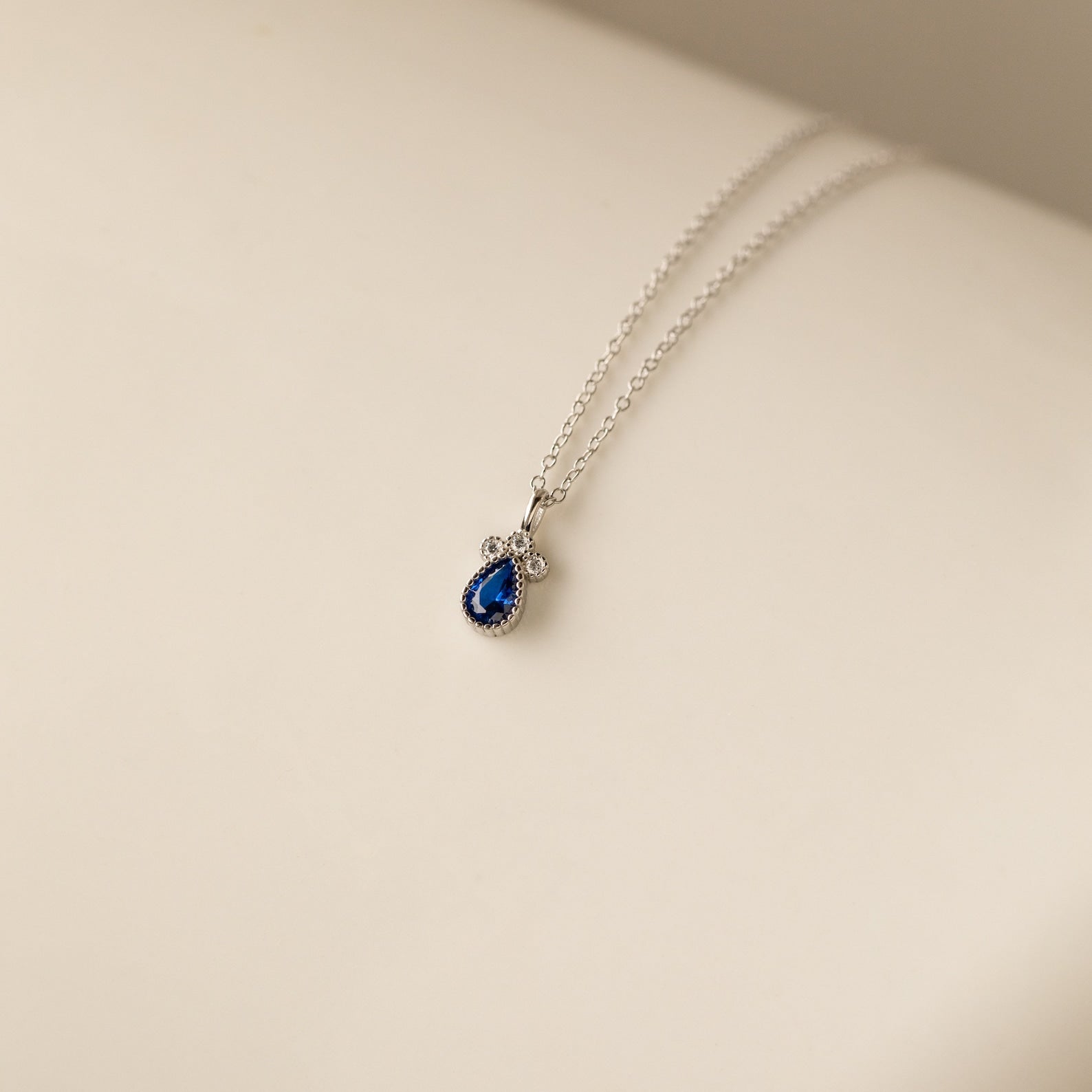 Lorelei Sapphire Teardrop Necklace with a blue sapphire gemstone pendant and delicate bow, elegantly displayed on a light cream background.