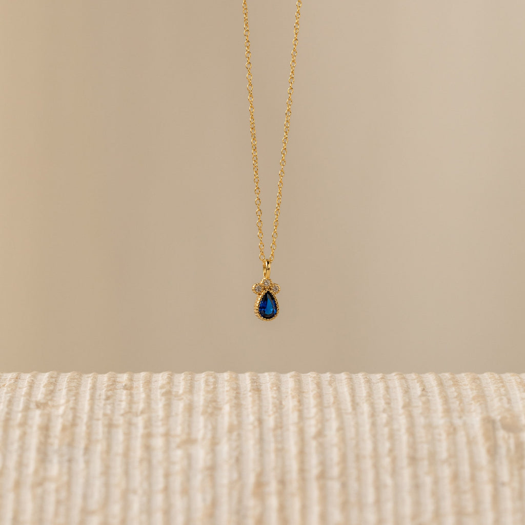 The Lorelei Sapphire Teardrop Necklace features a gold chain and a teardrop sapphire pendant, offering an elegant birthstone jewelry option that shines beautifully against any neutral background.