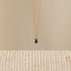 The Lorelei Sapphire Teardrop Necklace features a gold chain and a teardrop sapphire pendant, offering an elegant birthstone jewelry option that shines beautifully against any neutral background.