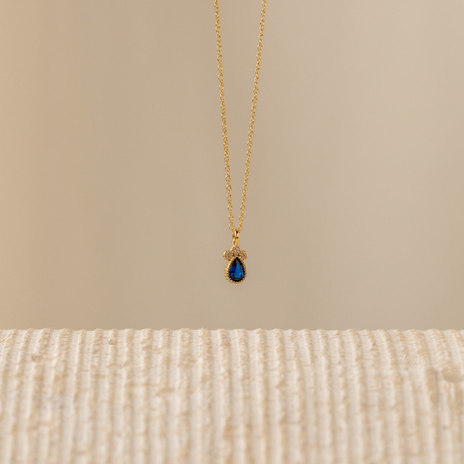 The Lorelei Sapphire Teardrop Necklace features a gold chain and a teardrop sapphire pendant, offering an elegant birthstone jewelry option that shines beautifully against any neutral background.