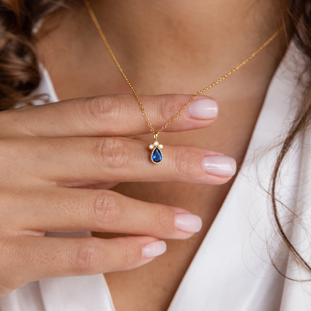 A hand displays the Lorelei Sapphire Teardrop Necklace, its rich blue gemstone pendant elegantly resting over a white blouse.