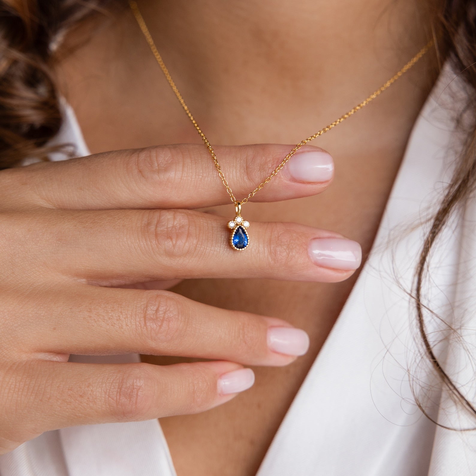 A hand displays the Lorelei Sapphire Teardrop Necklace, its rich blue gemstone pendant elegantly resting over a white blouse.