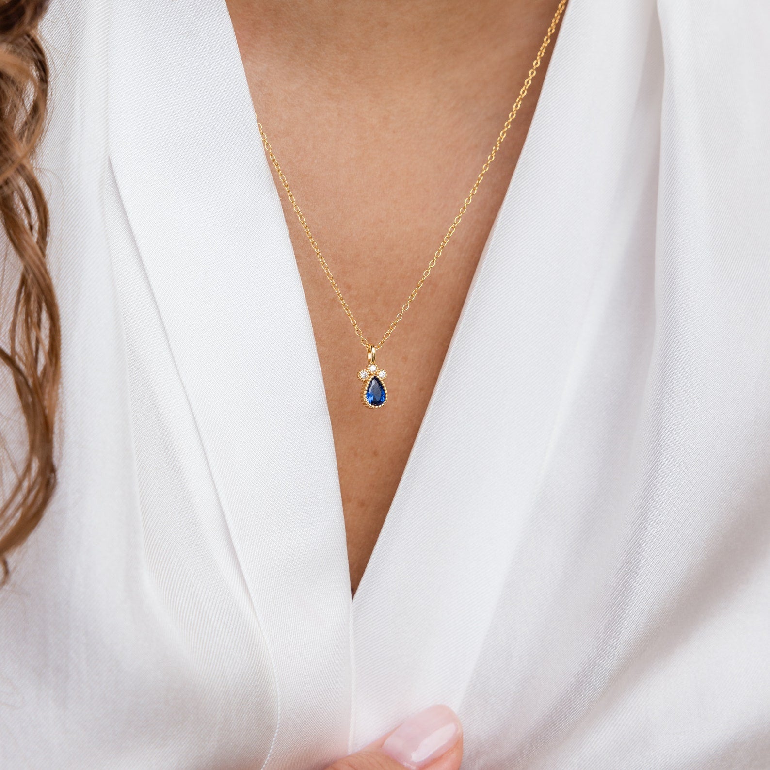 The Lorelei Sapphire Teardrop Necklace, featuring a blue pendant, elegantly drapes over a white blouse, enhancing the allure of this birthstone accessory.