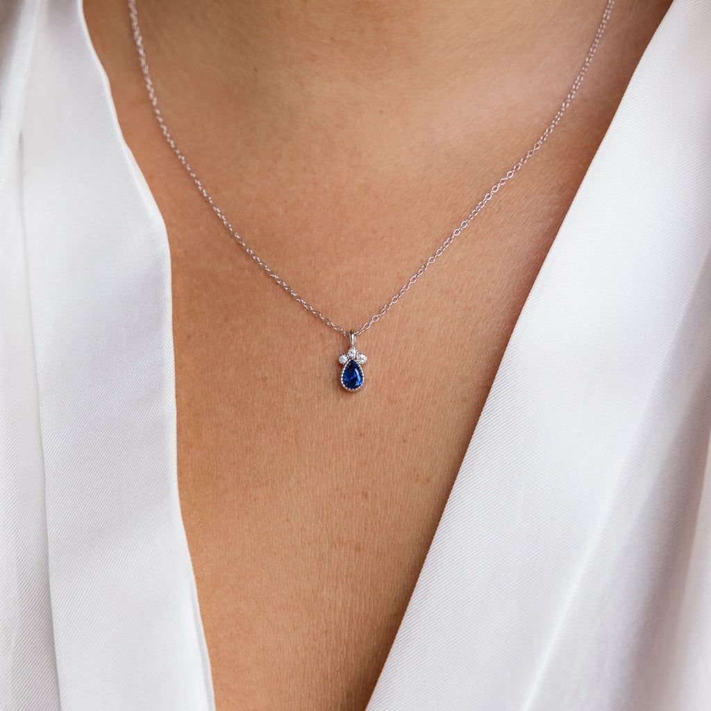 A close-up of the Lorelei Sapphire Teardrop Necklace, with its silver chain and sparkling blue pendant, elegantly showcased over a white blouse.