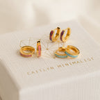 Four pairs of colorful Pastel Opal Inlay Huggies, featuring opal hoop and huggie earrings, displayed on a textured white Caitlyn Minimalist box.