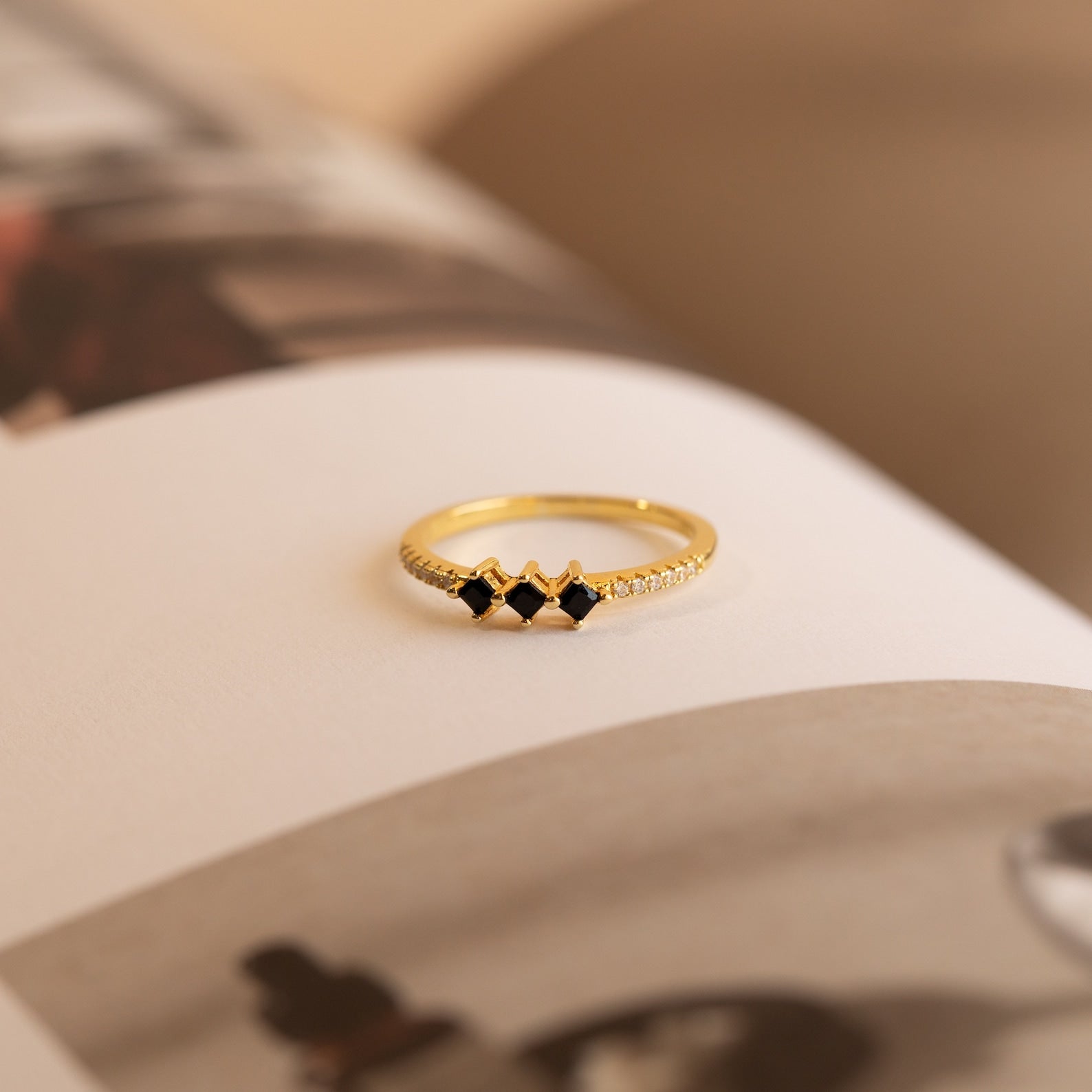 The Helena Black Diamond Ring features three black square stones and small clear accents, resting elegantly on an open book—perfect for adding an edgy, professional touch.