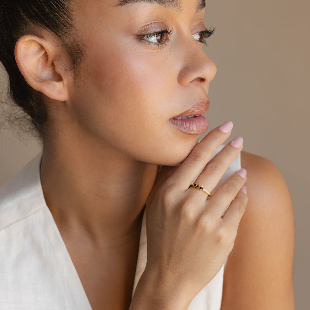 A woman with neutral makeup gazes sideways, her hand near her face showcasing the Helena Black Diamond Ring, wearing a light sleeveless top against a beige background.