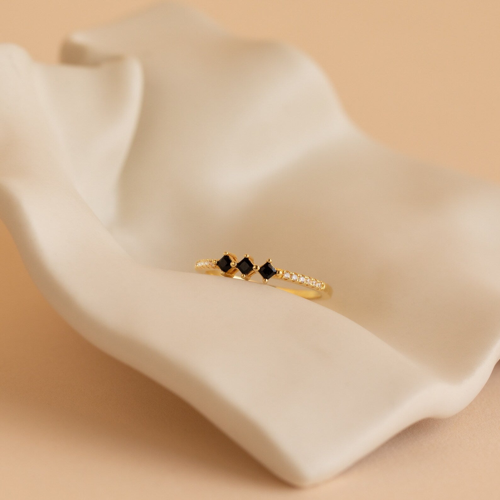 The Helena Black Diamond Ring, featuring black stones and small diamonds, rests on a sculpted beige ceramic surface, adding an edgy yet professional touch.