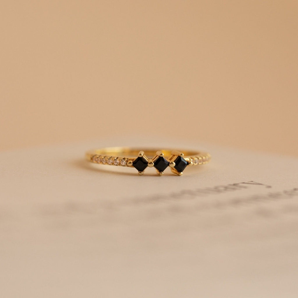 The Helena Black Diamond Ring is a delicate vintage gold ring with three black square stones and small clear gems on the band, set against a beige background for an edgy, professional look.