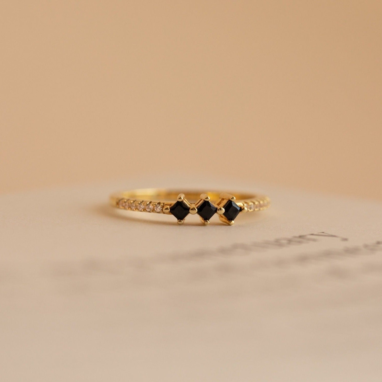 The Helena Black Diamond Ring is a delicate vintage gold ring with three black square stones and small clear gems on the band, set against a beige background for an edgy, professional look.