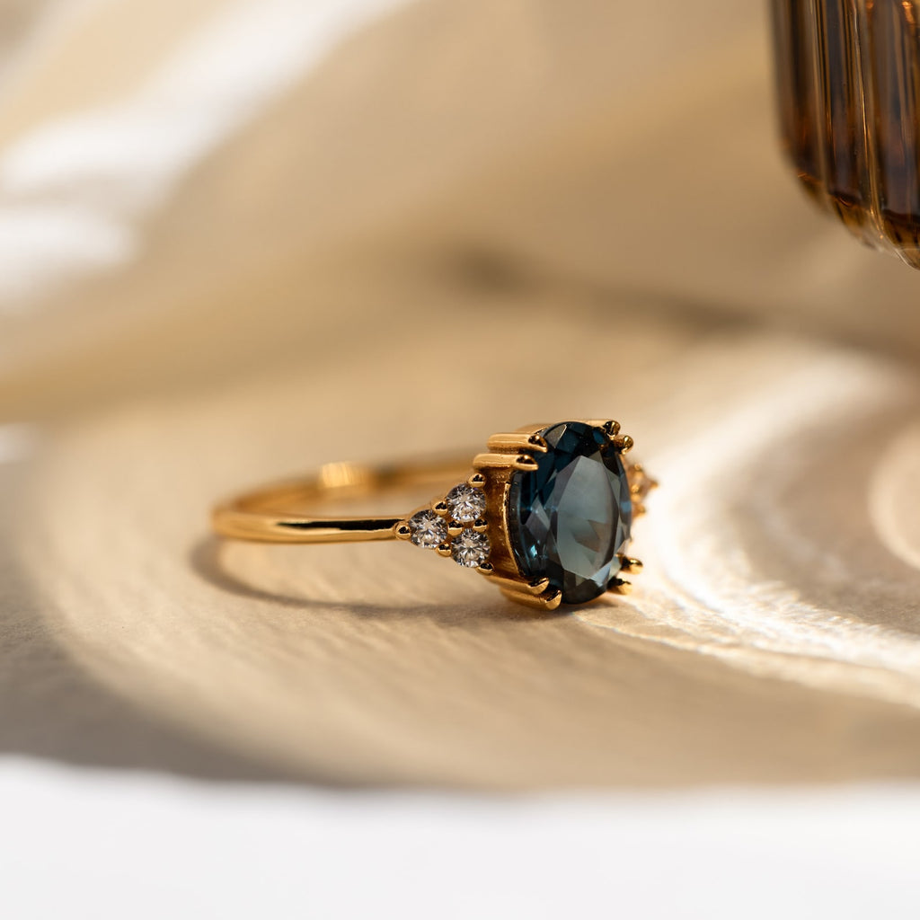 Elizabeth Blue Topaz Ring in 18K Gold featuring an oval blue topaz and small clear stones, shown on a light textured surface—a vintage boho-inspired design.