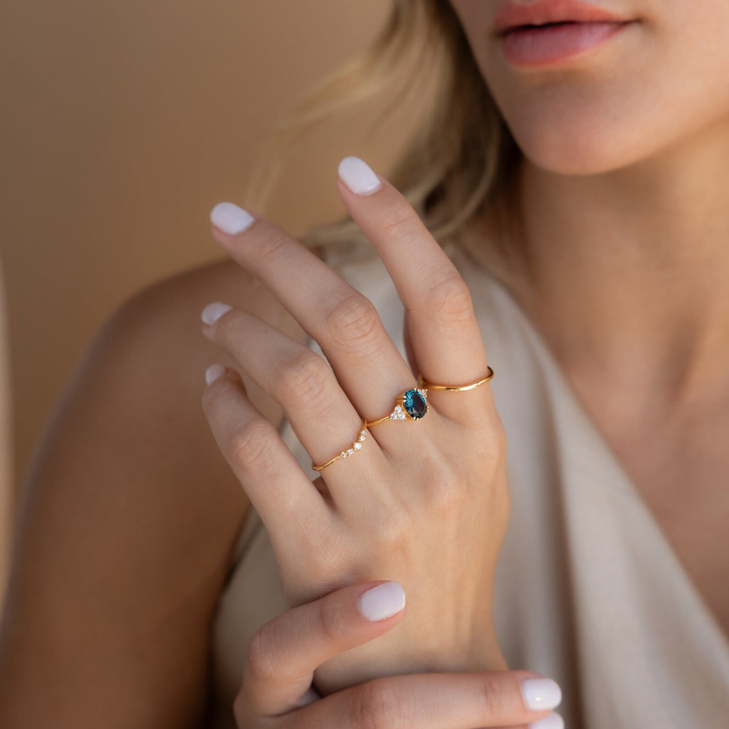 Close-up of a woman's hand wearing gold rings, including the Elizabeth Blue Topaz Ring in 18K Gold, featuring a vibrant blue gemstone, set against a neutral background.