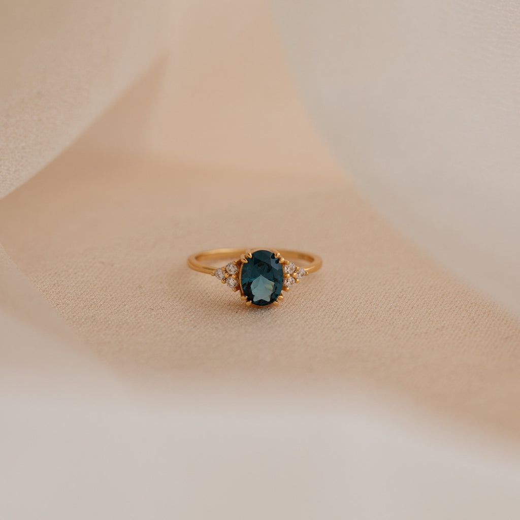 Elizabeth Blue Topaz Ring in 18K Gold, featuring an oval dark blue gemstone and small clear stones, showcased on a soft beige fabric background.