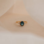 Elizabeth Blue Topaz Ring in 18K Gold, featuring an oval dark blue gemstone and small clear stones, showcased on a soft beige fabric background.