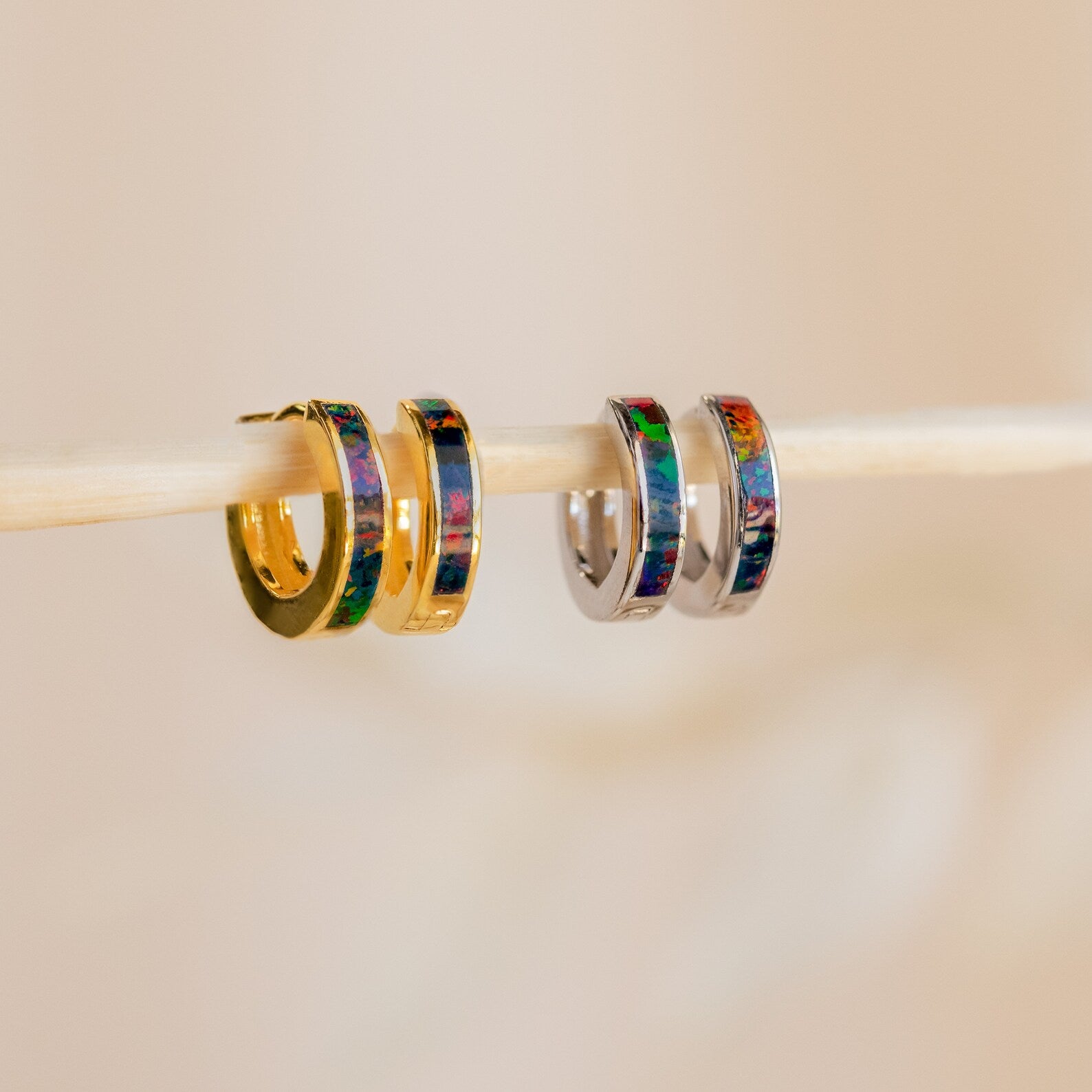 Two pairs of Black Opal Inlay Huggies—featuring gold and silver hoops with colorful inlays—hang on a stick.