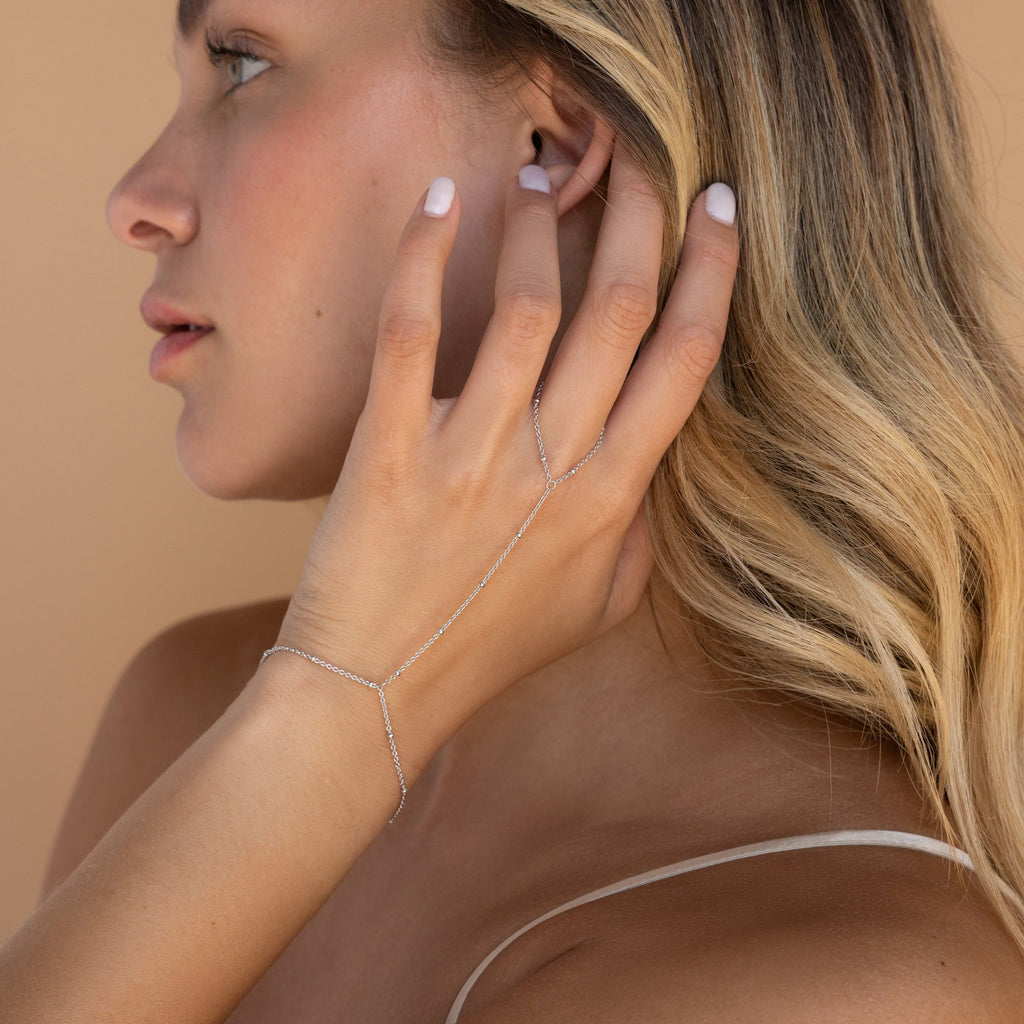 A blonde woman touches her ear, highlighting the Beaded Hand Chain Bracelet in Sterling Silver—a delicate statement piece—set against a beige background.