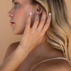 A blonde woman touches her ear, highlighting the Beaded Hand Chain Bracelet in Sterling Silver—a delicate statement piece—set against a beige background.
