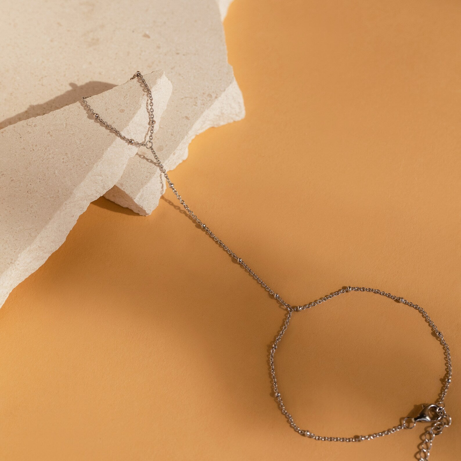 A delicate Beaded Hand Chain Bracelet rests on a beige stone against a warm tan background, offering an understated statement piece for any jewelry lover.