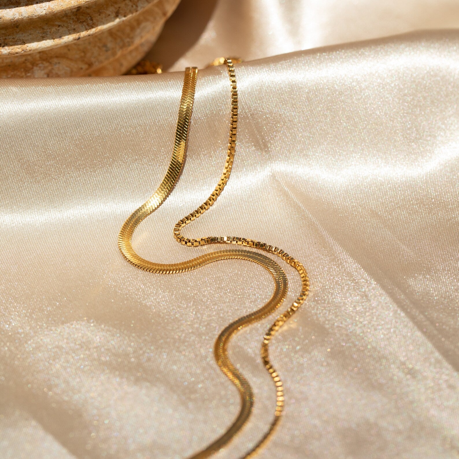 The Herringbone Duo Chain Necklace, a minimalist gold piece, is shown resting elegantly on shiny cream satin fabric.