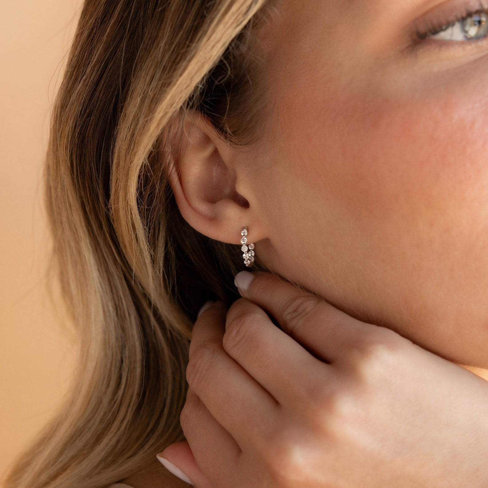A woman’s ear adorned with Inside Out Diamond Huggies, sparkling hoops; her hand softly rests on her neck.