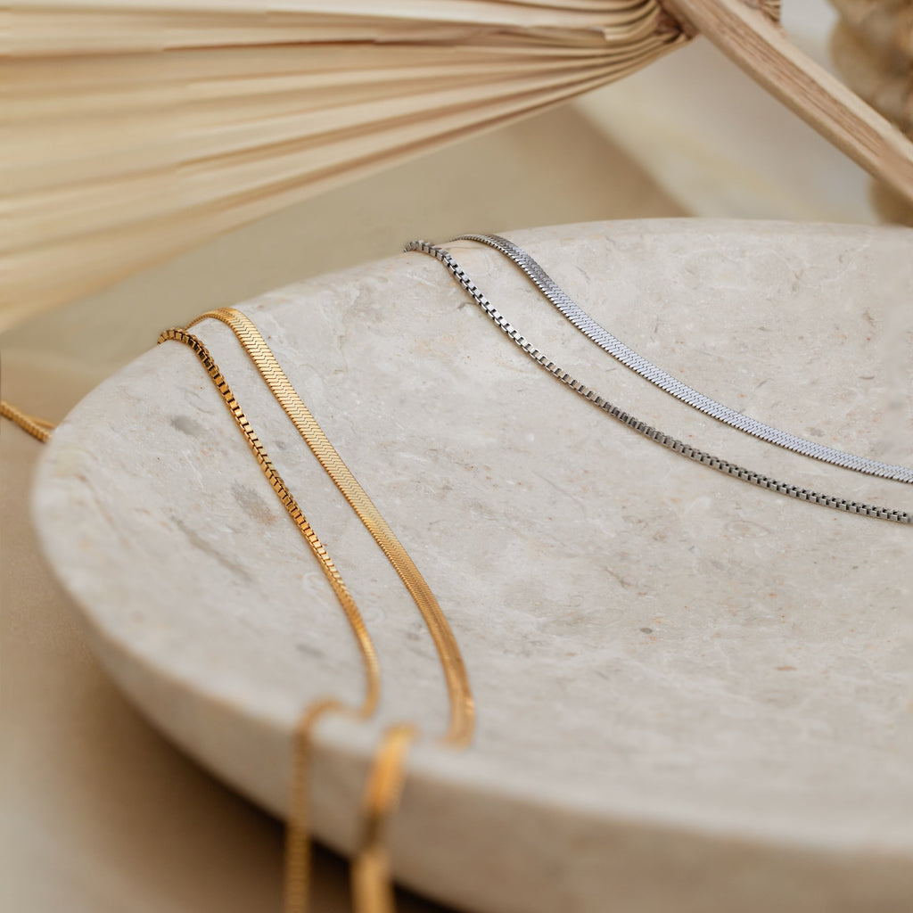 The Herringbone Duo Chain Necklace in gold and silver rests on a round, beige stone dish with a neutral backdrop—ideal for anyone who loves minimalist necklace styles.