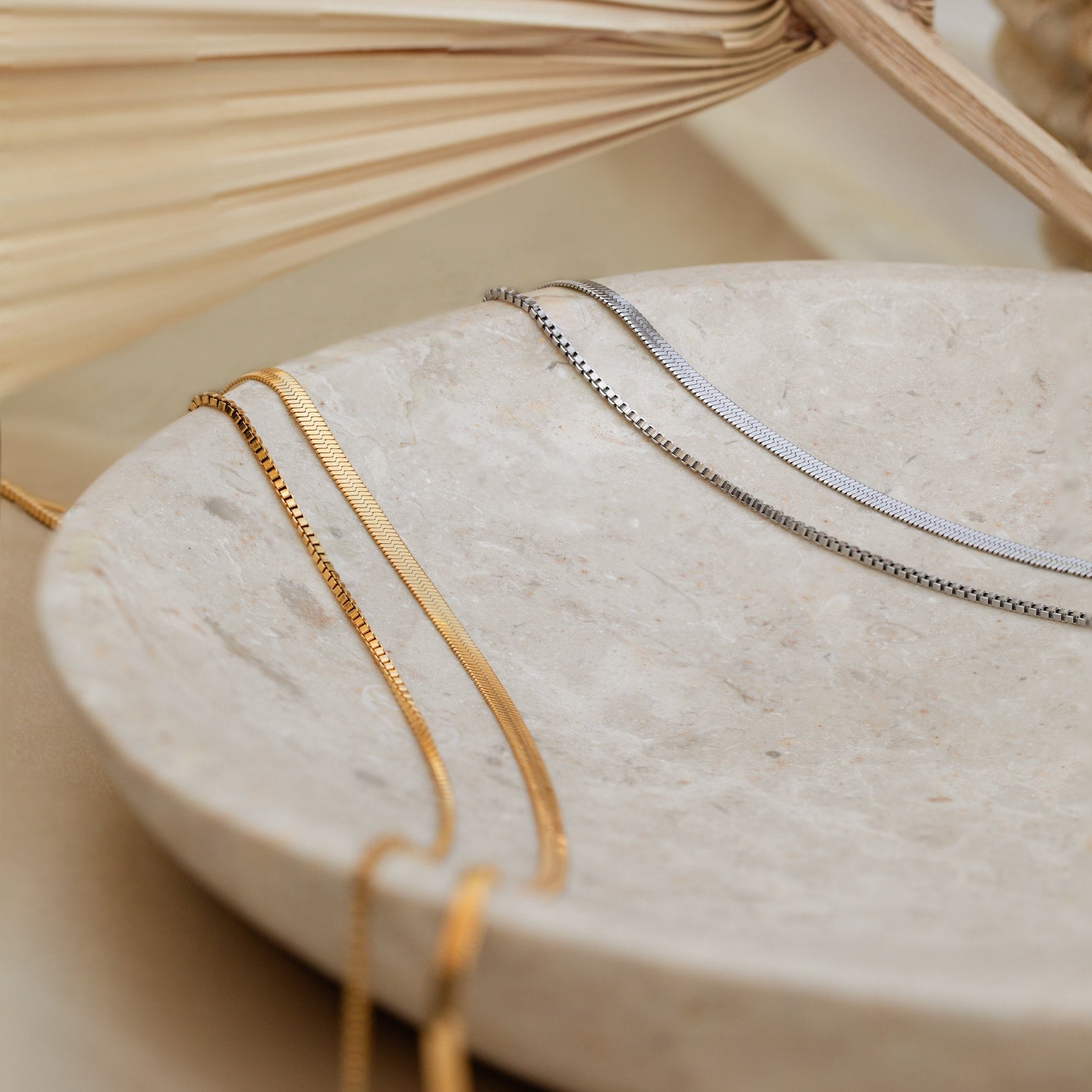 The Herringbone Duo Chain Necklace in gold and silver rests on a round, beige stone dish with a neutral backdrop—ideal for anyone who loves minimalist necklace styles.