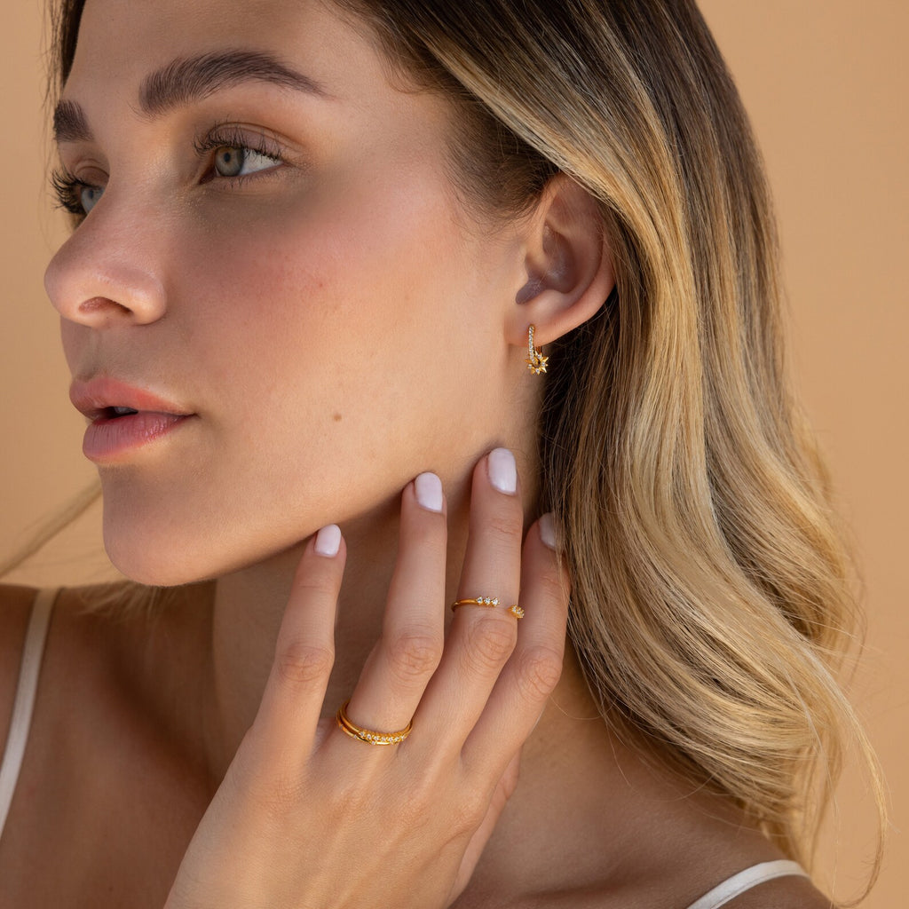 A woman with wavy blonde hair wears Pave Sun Huggies gold earrings, touching her face and looking left, against a beige background.